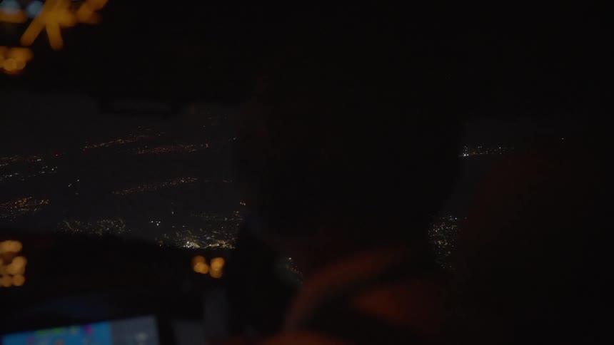 From behind the pilot in a dark cockpit, the view shows a vast, illuminated city sprawling below during a nighttime flight