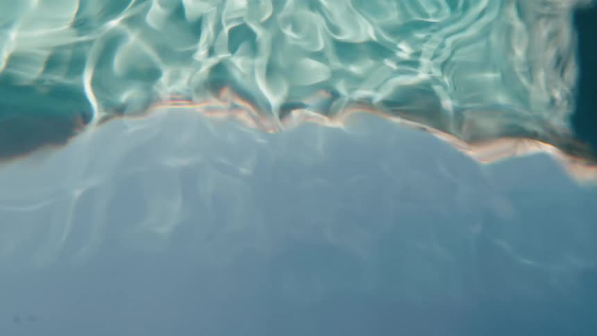An underwater view of a swimmer's distorted silhouette moving across the shimmering, sunlit surface of a pool