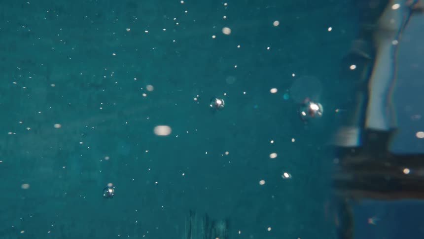 An abstract underwater close-up of shiny air bubbles rising through deep teal water in slow motion