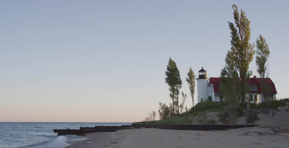A picturesque lighthouse stands on a sandy shore as gentle waves lap the beach at sunset