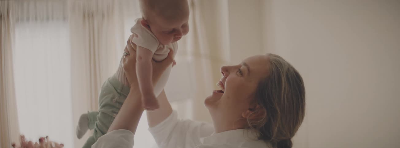 A smiling mother playfully lifts her sleepy newborn baby into the air and gives them a tender kiss in a softly lit room