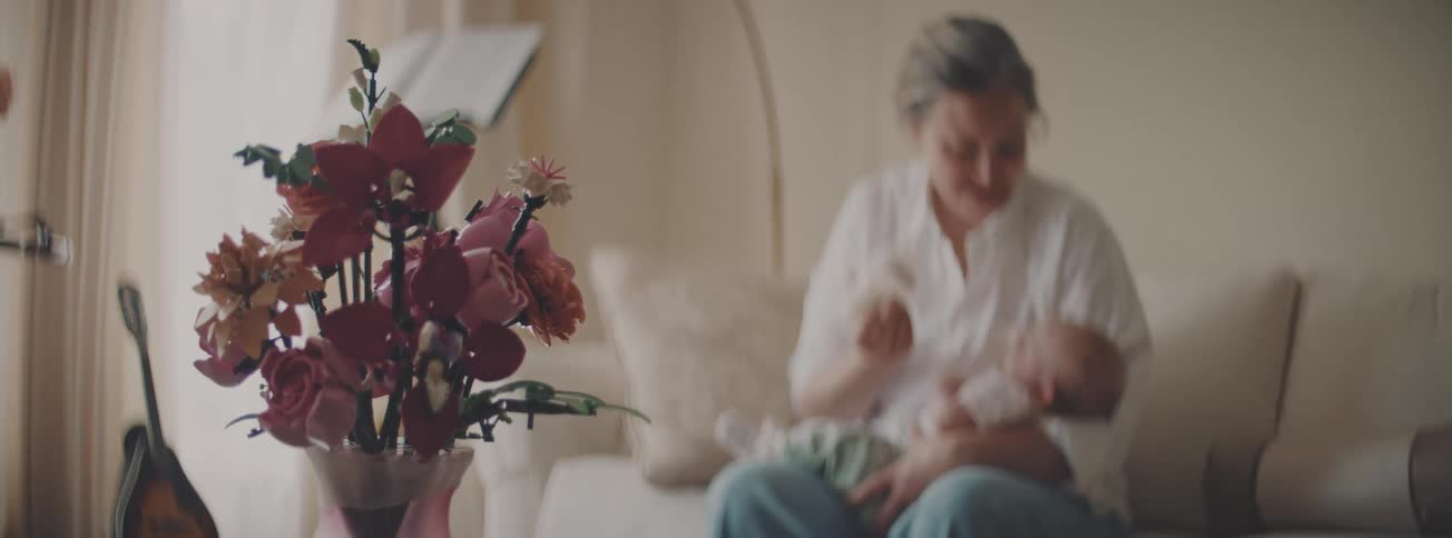 A mother bottle-feeds her newborn baby in a softly lit living room, with a LEGO flower bouquet in the foreground