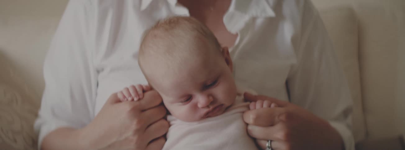 A newborn baby with scrunched-up eyes slowly falls asleep while being held in their mother's arms
