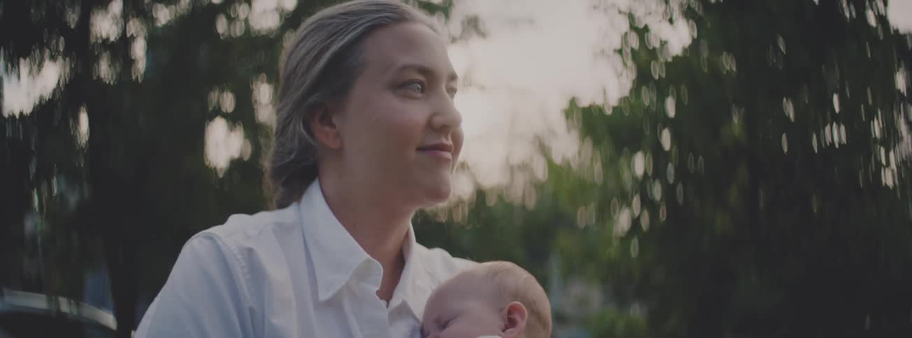 A happy young mother in a white shirt gently holds her sleeping baby in her arms while looking off peacefully into the distance outdoors