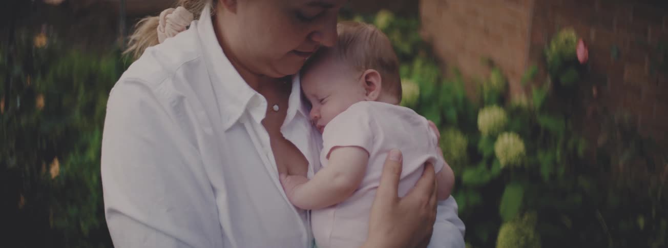 A mother gently kisses her sleeping newborn baby's head while holding her in a lush garden