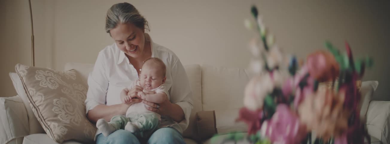 A loving grandmother smiles as she holds her newborn grandchild while sitting on a couch in a cozy living room
