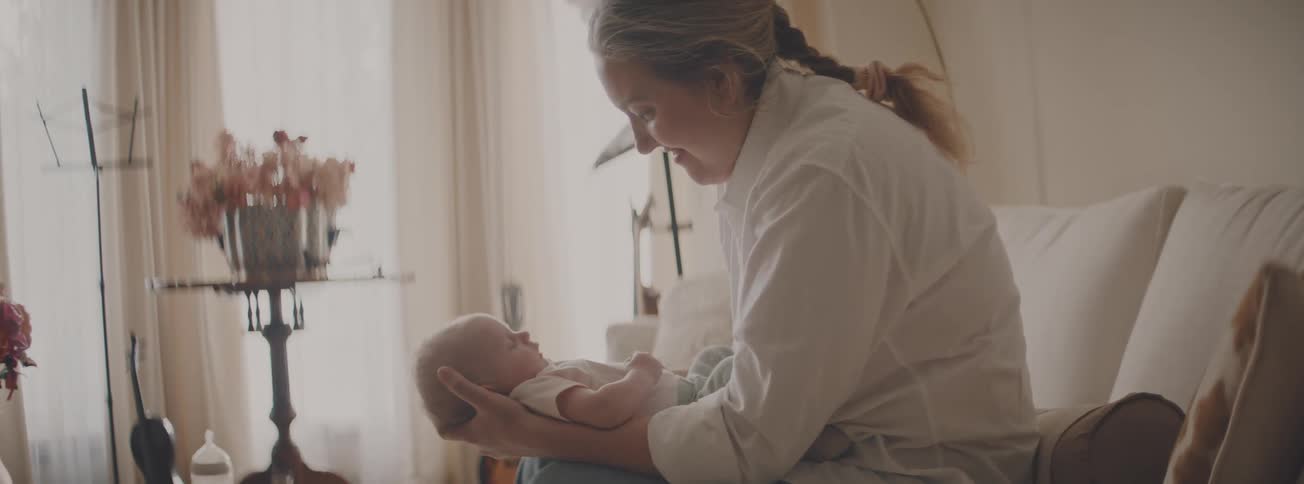 A loving mother smiles as she holds her newborn baby in her lap in a bright and cozy living room