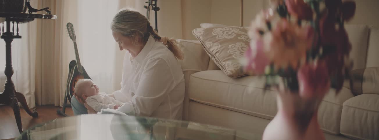 A loving grandmother holds and smiles at her newborn grandchild on the floor of a sunlit living room
