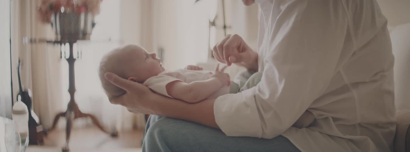 A mother gently holds her newborn baby in her lap, tenderly touching its nose and chest in a softly lit room