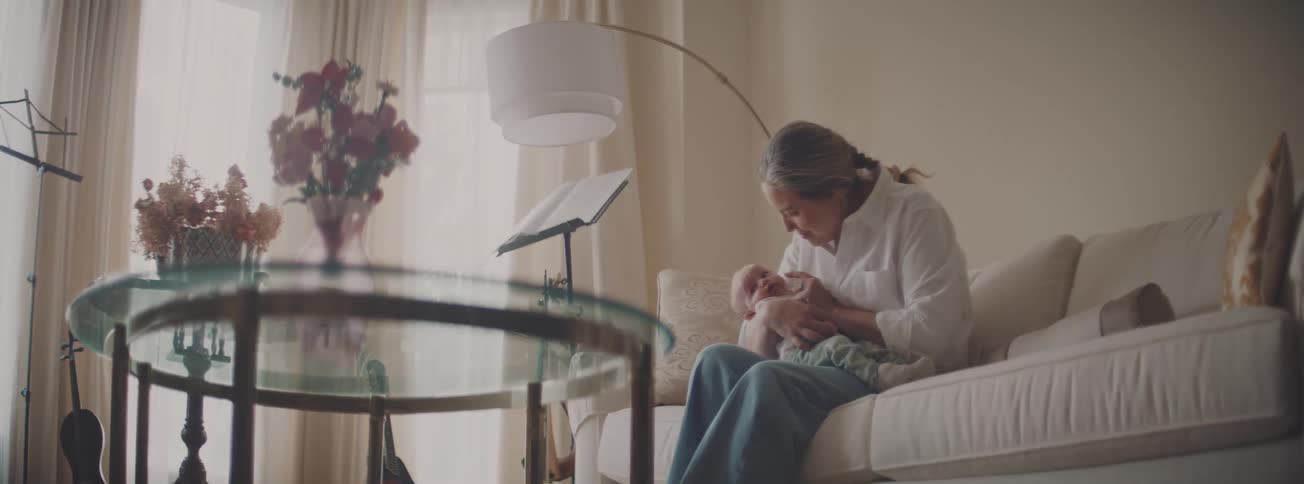 A loving woman sits on a sofa and gently holds a newborn baby in her lap in a warm, sunlit living room