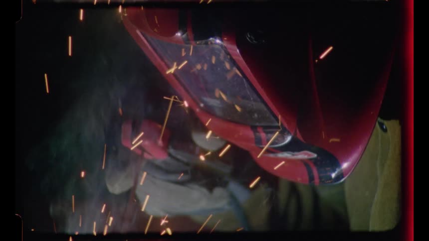 A high-angle close-up of a person in a red welding mask working on metal as sparks and smoke fly, captured on vintage film