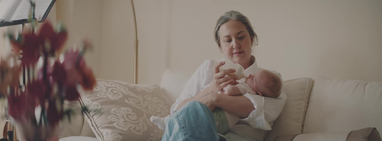 A gray-haired mother tenderly feeds her newborn baby with a bottle while sitting on a comfortable sofa at home
