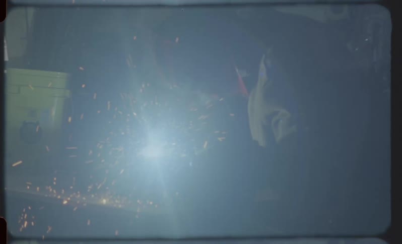 A welder in a dark workshop creates a shower of sparks in this hazy, vintage-style film footage with a rainbow lens flare