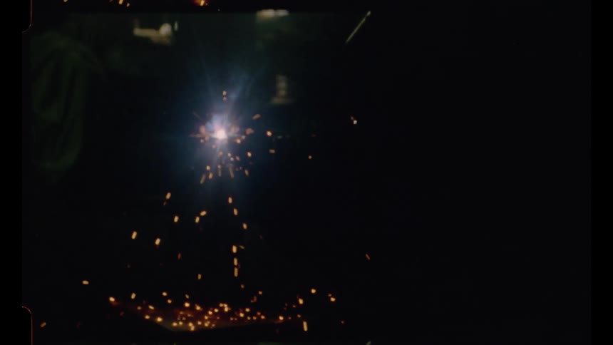 Vintage 8mm footage captures a welder at work in a dimly lit shop as bright sparks fly from the torch