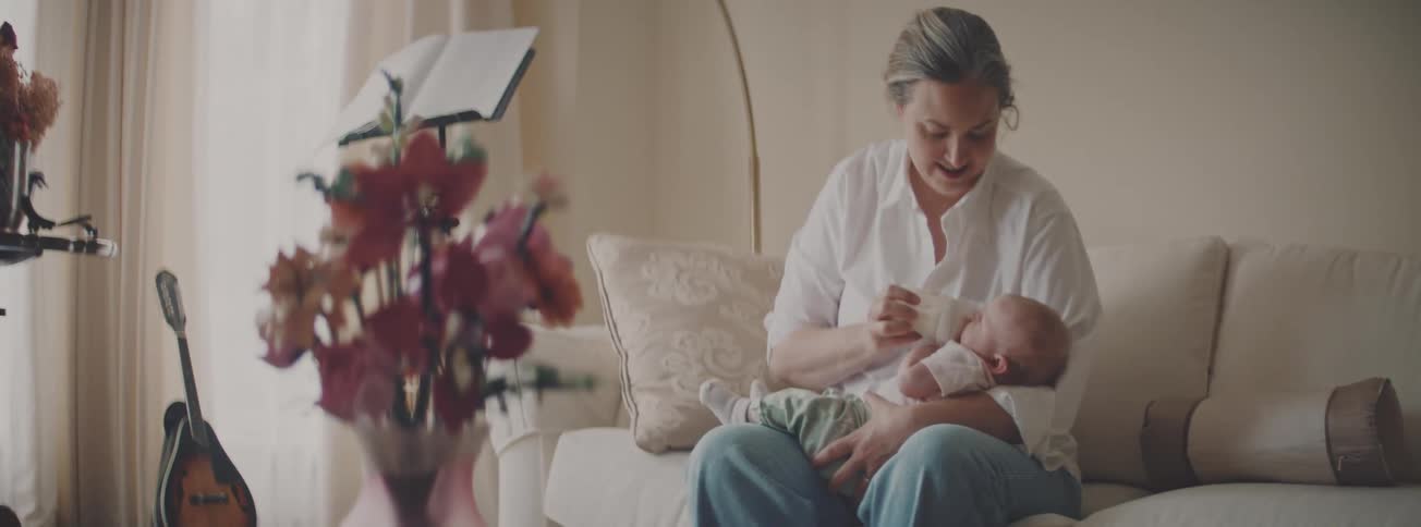 A mother tenderly bottle-feeds her newborn baby while sitting on a cozy sofa in a softly lit living room