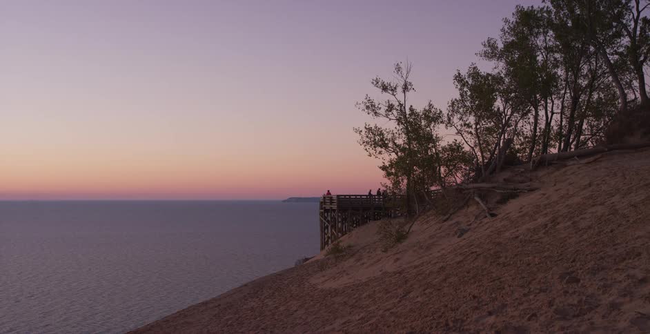 A tranquil view from a sandy dune shows a wooden lookout over the calm ocean during a beautiful purple and pink sunset