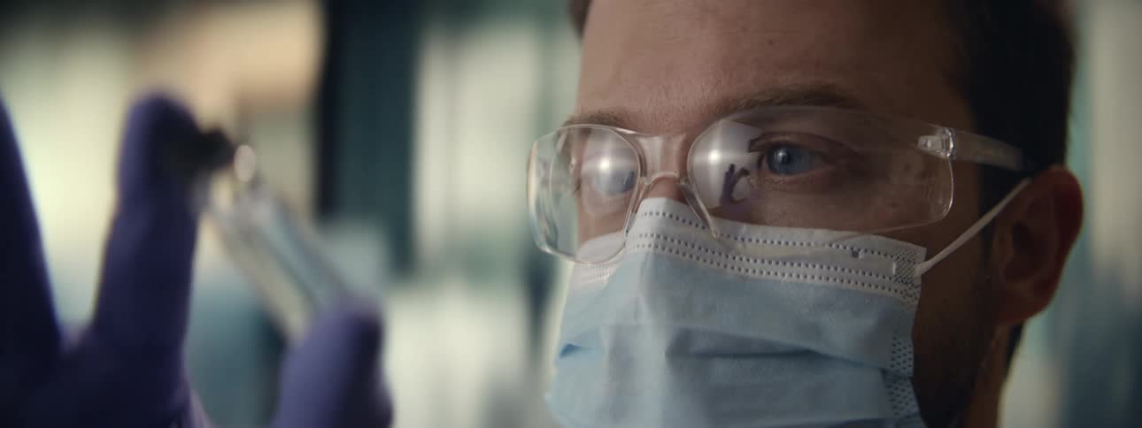 Close up shot of a determined scientist with blue eyes wearing a mask and safety goggles examining a vial in a lab