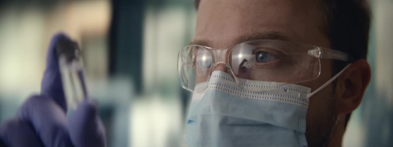 A scientist with blue eyes wearing a mask and safety goggles intently studies a sample in a test tube in his laboratory