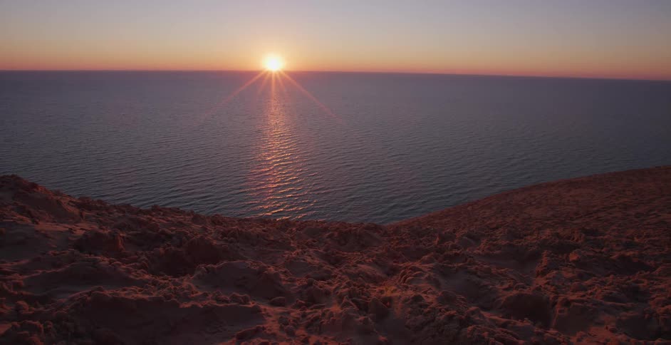 The sun sets over the ocean, casting a warm glow on the water and the sandy cliff in the foreground