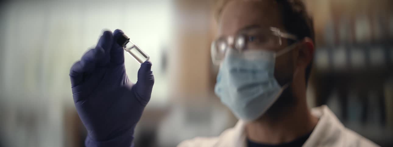 A scientist in a lab coat, mask, and gloves carefully examines a small glass vial containing a clear liquid sample