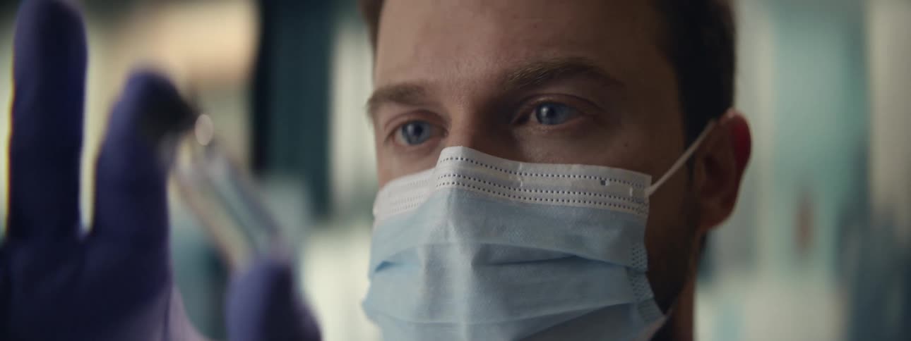 A close-up of a scientist with blue eyes wearing a mask and gloves, intently examining a small glass vial in a laboratory