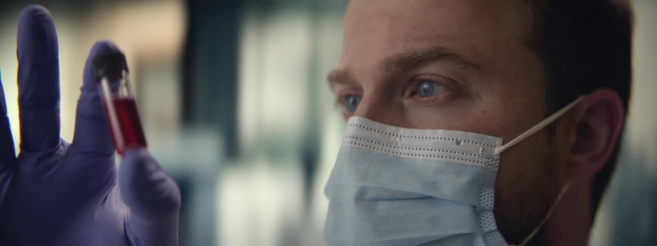 A focused male scientist in a face mask and gloves carefully examines a vial containing a red blood sample in a laboratory