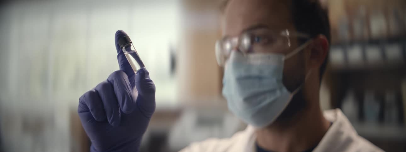 A focused male scientist wearing a mask and gloves carefully examines a small glass vial in a research laboratory