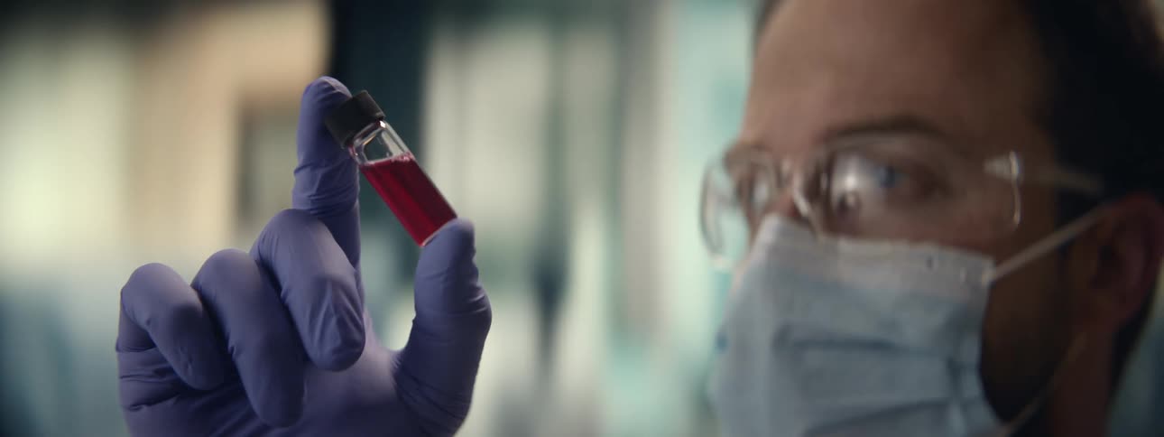 A scientist wearing protective gloves, mask, and glasses closely examines a vial of red liquid in a laboratory setting