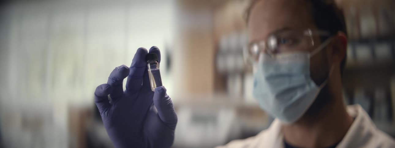 A focused scientist wearing protective gear carefully inspects a small glass vial containing a clear liquid in a laboratory