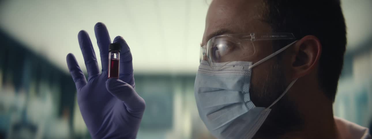 A male scientist wearing a mask and safety goggles holds up a vial containing a red liquid to examine it in a lab