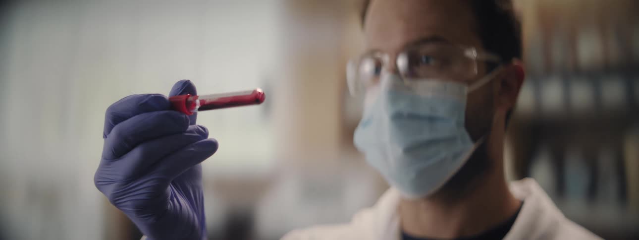 A scientist in a lab coat, mask, and gloves carefully inspects a vial of blood