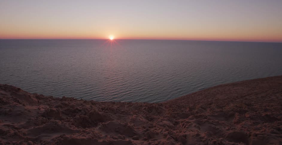 A peaceful sunset casts a golden glow over the calm ocean from the top of a sandy dune