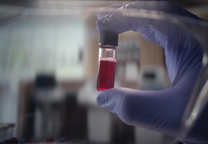 A gloved hand holds up a small glass vial containing a bubbling red liquid for inspection inside a laboratory