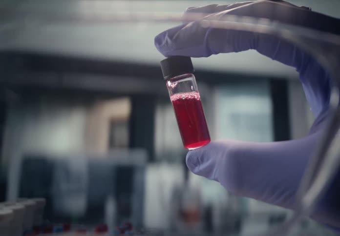 A scientist in a sterile lab holds up a glass vial containing a red liquid sample for analysis