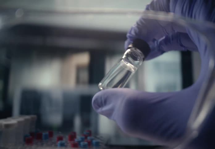 A scientist wearing a protective glove carefully examines a small vial containing a clear liquid in a sterile laboratory setting