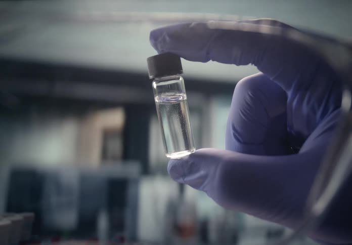 A scientist in a lab holds up and shakes a glass vial containing a clear liquid creating bubbles
