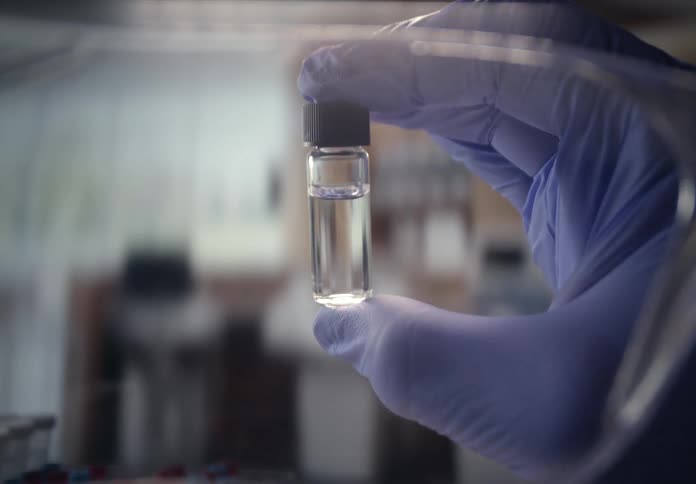 A scientist in a blue glove holds up a small glass vial of clear liquid for inspection in a sterile laboratory setting