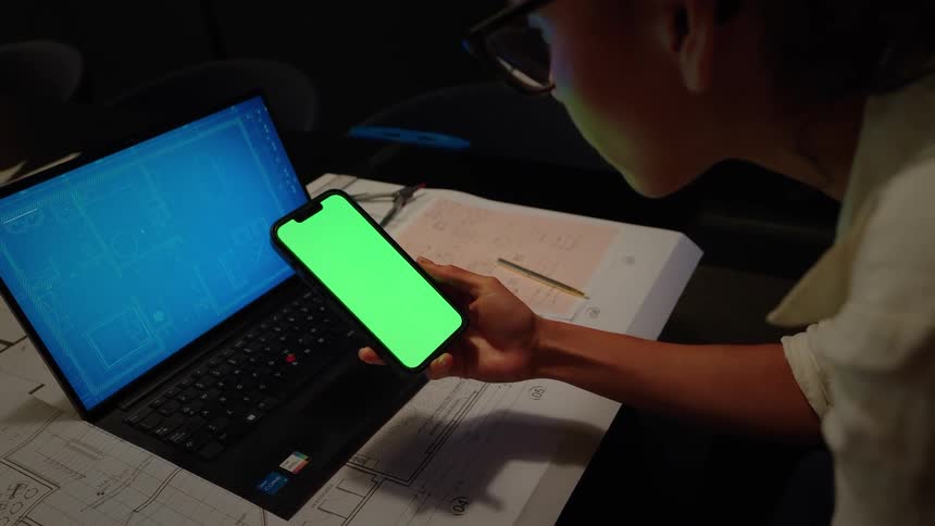 Over the shoulder view of an architect looking at a green screen smartphone while working on blueprints at their desk late at night