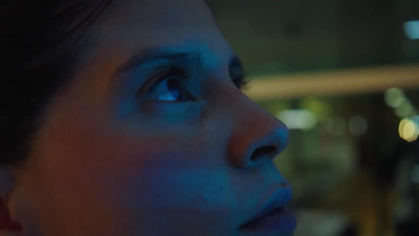 A thoughtful young woman watches something off-screen in a cinematic close-up, her face illuminated by a moody blue light