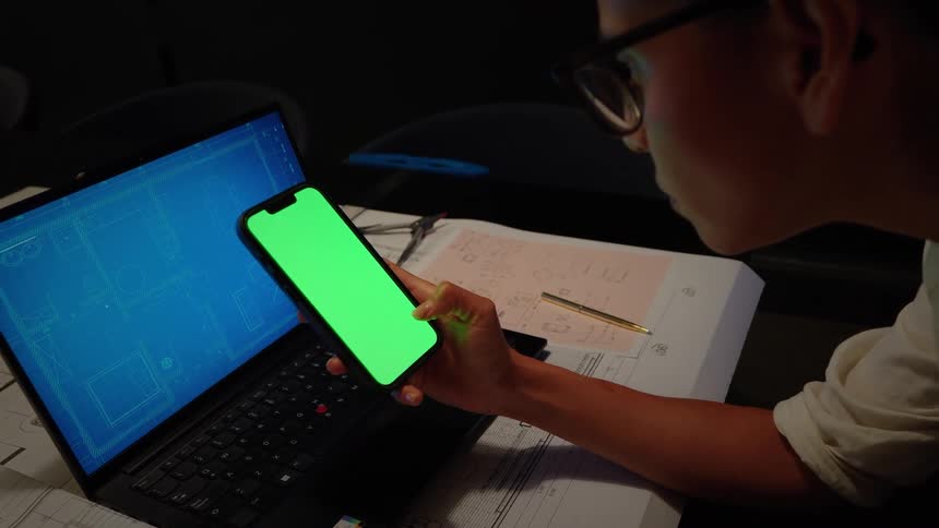 An architect works late at night in a dark office, viewing blueprints on her laptop and using a smartphone with a green screen