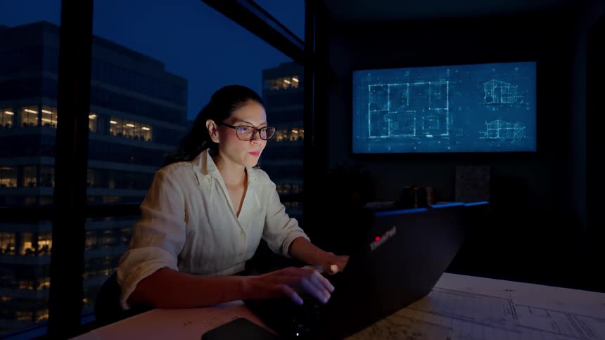 A professional female architect works late at night on a laptop in a modern office with a view of the city skyline