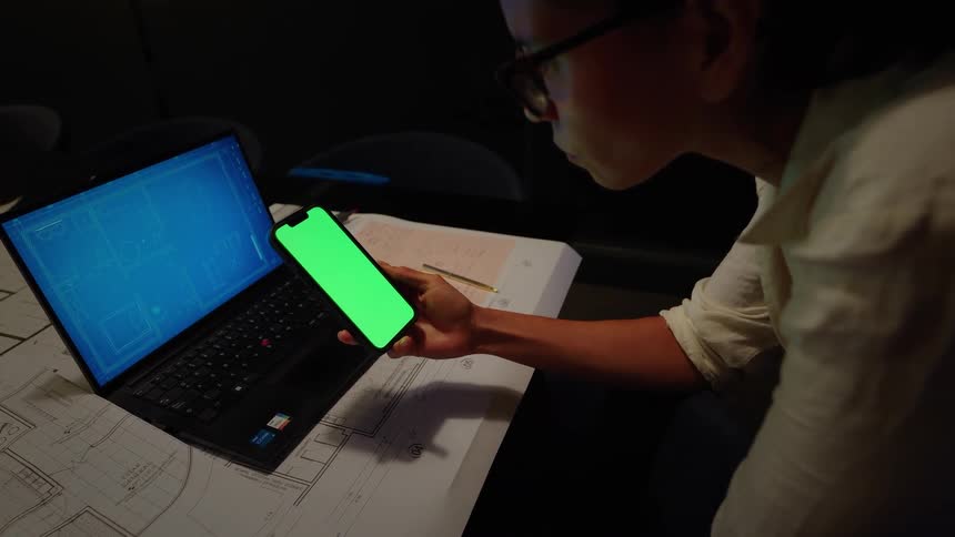 A female architect works late in a dark office, using a green-screen smartphone with blueprints on her laptop and desk