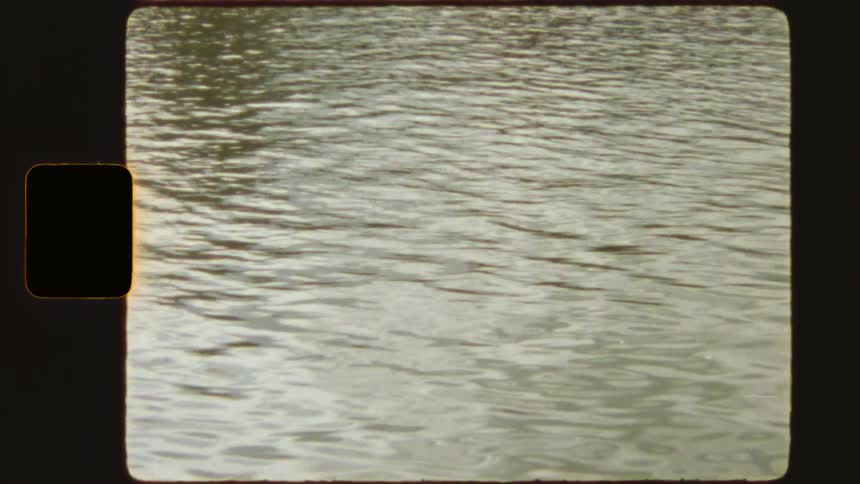 Vintage Super 8 film captures a view from a boat on a city river, revealing an old industrial building with graffiti on the bank