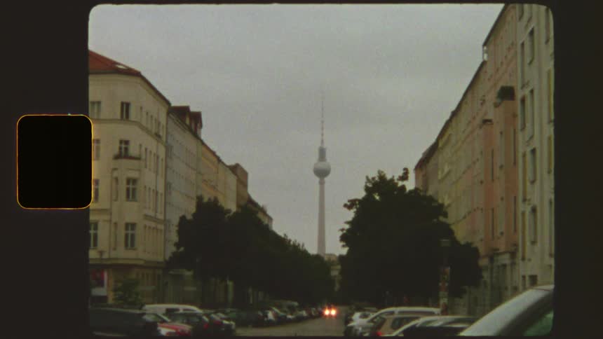 Vintage Super 8mm film footage zooms out from the Berlin TV Tower to reveal a city street lined with cars and apartment buildings on a cloudy day