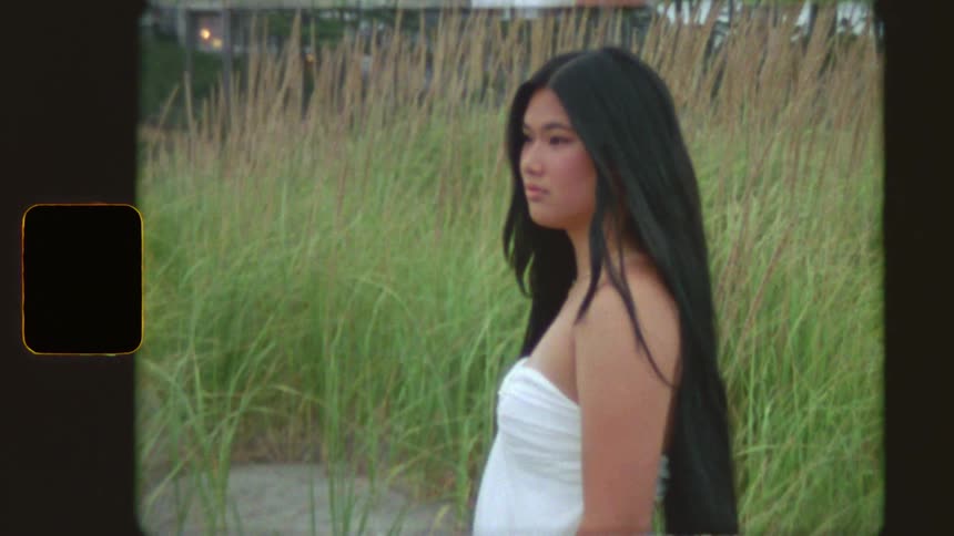 Vintage film captures a young woman with long black hair standing in a field of tall grass before she turns to smile at the camera