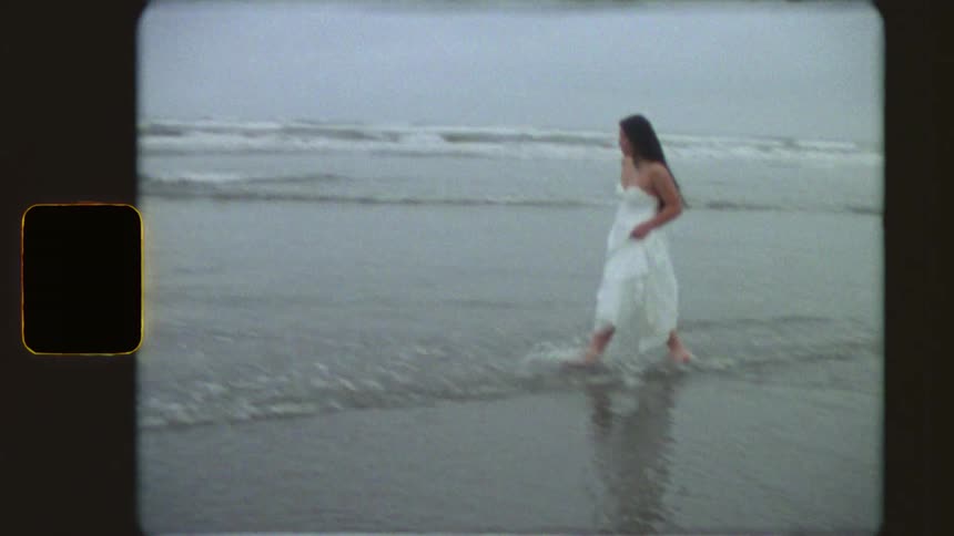 Vintage Super 8 footage captures a woman with long dark hair in a white dress running barefoot along the shoreline before walking and splashing in the waves