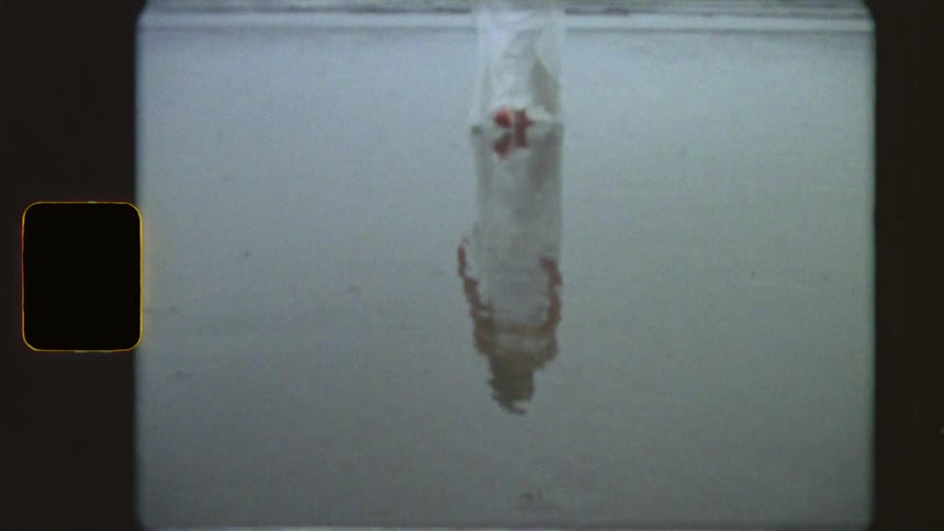 A woman in a long white dress walks barefoot along the water's edge on a foggy beach, her reflection captured in the wet sand in this vintage 8mm footage