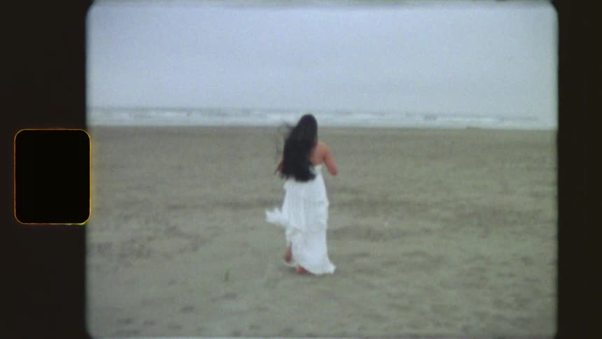 A woman in a flowing white dress runs barefoot across a wide sandy beach towards the ocean in a dreamy vintage Super 8 film