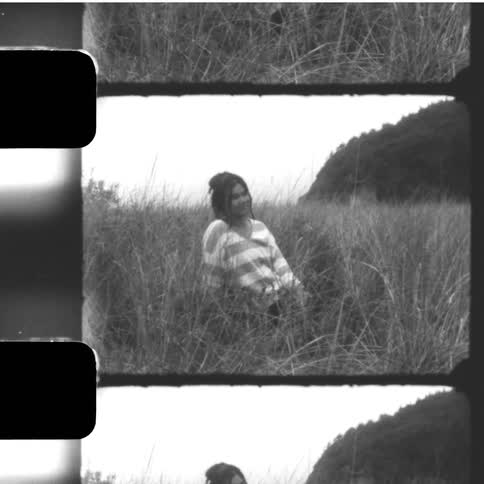 Vintage black and white filmstrip footage of a woman sitting in a field of tall grass, turning to smile at the camera