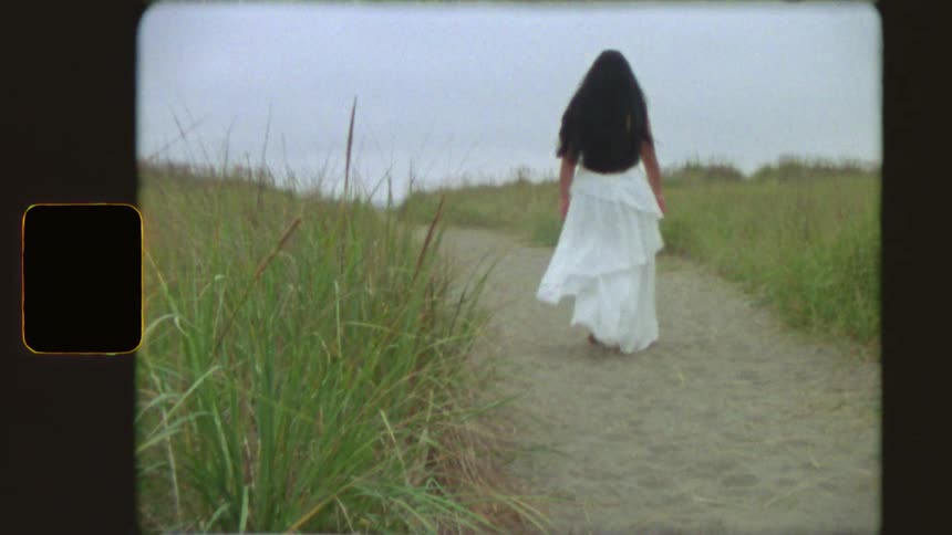 A woman in a flowing white dress walks away down a sandy path through tall beach grass in this vintage Super 8 film